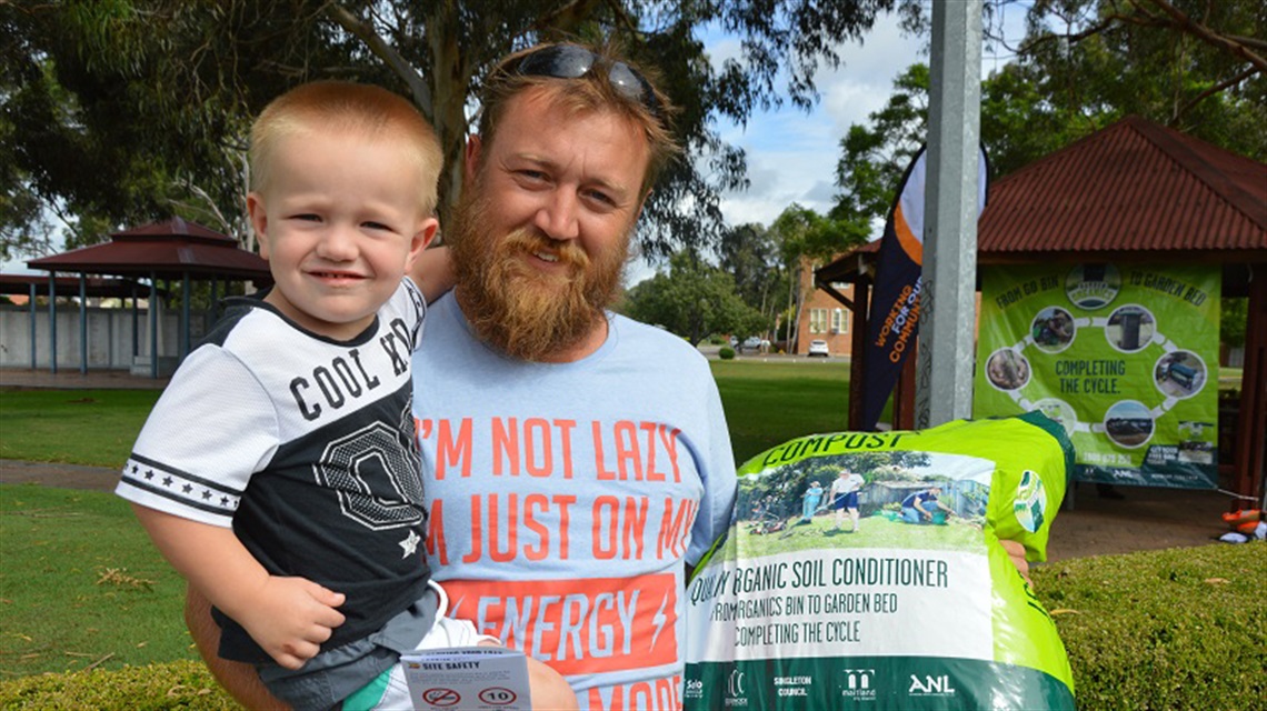 Photo of a man at a compost giveaway event holding a toddler and a bag of compost
