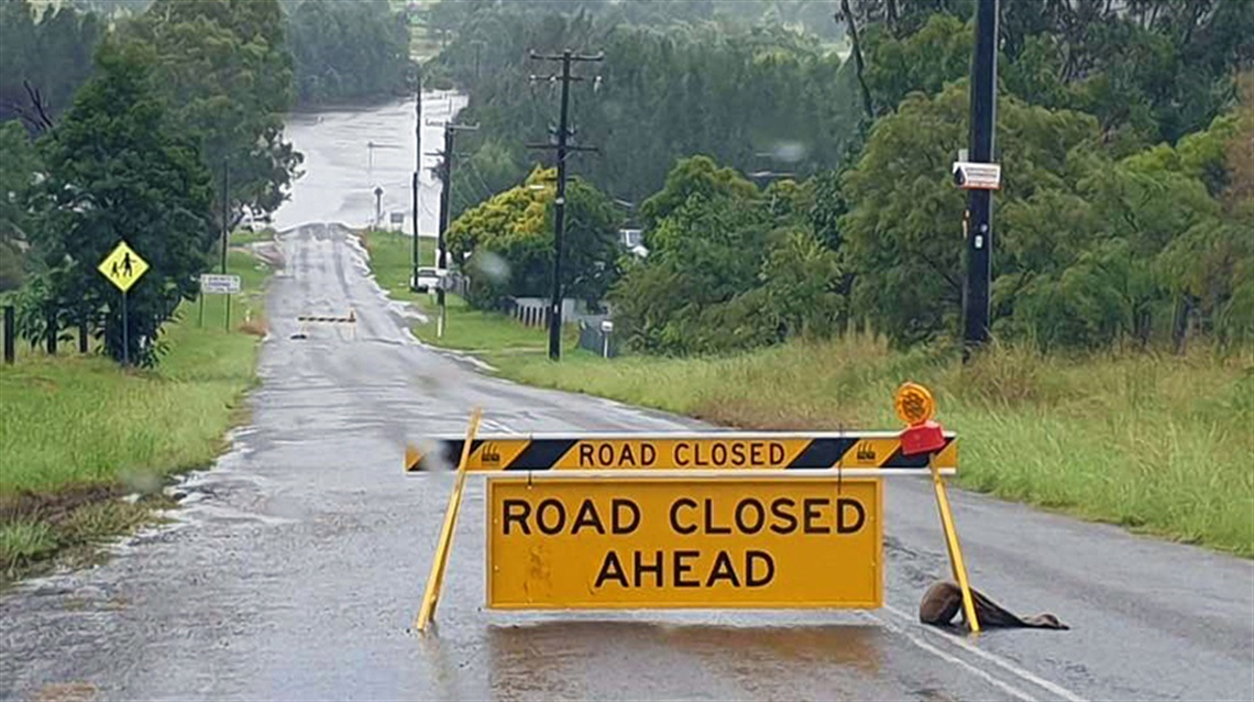 Photo of Congewai Road with a yellow and black ROAD CLOSED AHEAD sign in the foreground and a flooded road in the background